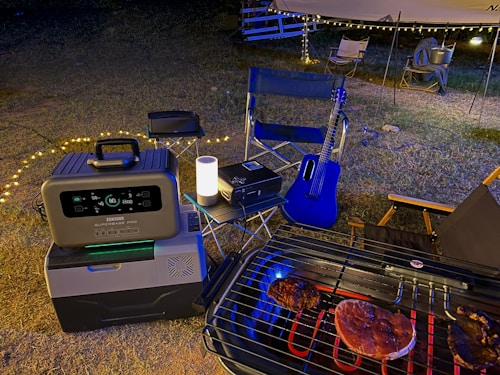 A nighttime outdoor scene featuring a portable power station and cooler on the ground next to a folding chair. A grill with meat is situated in the foreground. A blue acoustic guitar leans against a chair under a canopy with string lights, creating a cozy atmosphere.