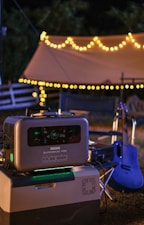 An outdoor setting with string lights creating a warm and cozy atmosphere. A portable power station with a digital display showing charge details sits prominently in the foreground on a larger device. In the background, a guitar is leaning against a chair near a tent.