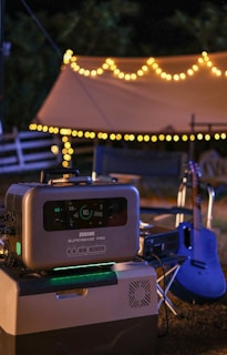 An outdoor setting with string lights creating a warm and cozy atmosphere. A portable power station with a digital display showing charge details sits prominently in the foreground on a larger device. In the background, a guitar is leaning against a chair near a tent.