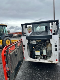 A construction vehicle with an open engine compartment revealing the engine and components. The vehicle is parked on a wet, asphalt surface with visible mud and dirt. In the background, there is another tractor vehicle with yellow wheels, and various industrial equipment and shipping containers can be seen.