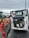 A construction vehicle with an open engine compartment revealing the engine and components. The vehicle is parked on a wet, asphalt surface with visible mud and dirt. In the background, there is another tractor vehicle with yellow wheels, and various industrial equipment and shipping containers can be seen.