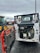 A construction vehicle with an open engine compartment revealing the engine and components. The vehicle is parked on a wet, asphalt surface with visible mud and dirt. In the background, there is another tractor vehicle with yellow wheels, and various industrial equipment and shipping containers can be seen.