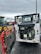 A construction vehicle with an open engine compartment revealing the engine and components. The vehicle is parked on a wet, asphalt surface with visible mud and dirt. In the background, there is another tractor vehicle with yellow wheels, and various industrial equipment and shipping containers can be seen.