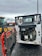 A construction vehicle with an open engine compartment revealing the engine and components. The vehicle is parked on a wet, asphalt surface with visible mud and dirt. In the background, there is another tractor vehicle with yellow wheels, and various industrial equipment and shipping containers can be seen.