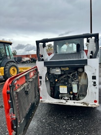 A construction vehicle with an open engine compartment revealing the engine and components. The vehicle is parked on a wet, asphalt surface with visible mud and dirt. In the background, there is another tractor vehicle with yellow wheels, and various industrial equipment and shipping containers can be seen.