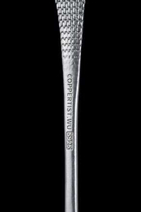 A close-up view of a silver metallic object with a textured surface. The object is inscribed with the text 'COPPERIST.WU S925'. The background is solid black, enhancing the focus on the metallic object.