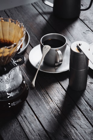 A rustic wooden table set with a French press, coffee grinder, and a cup of black coffee.