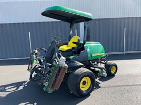 A green and yellow heavy-duty lawn mower is parked on an asphalt surface in front of a grey metal container. The mower has a canopy attached for shading and multiple cutting attachments on the front. It has large, rugged tires for stability and maneuverability.