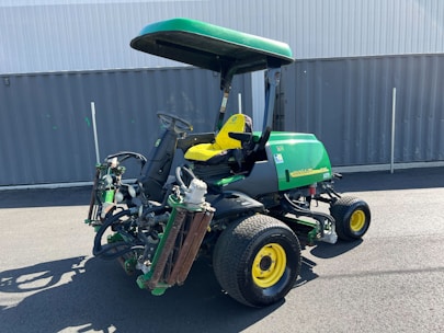 A green and yellow heavy-duty lawn mower is parked on an asphalt surface in front of a grey metal container. The mower has a canopy attached for shading and multiple cutting attachments on the front. It has large, rugged tires for stability and maneuverability.