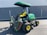 A green and yellow heavy-duty lawn mower is parked on an asphalt surface in front of a grey metal container. The mower has a canopy attached for shading and multiple cutting attachments on the front. It has large, rugged tires for stability and maneuverability.