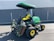 A green and yellow heavy-duty lawn mower is parked on an asphalt surface in front of a grey metal container. The mower has a canopy attached for shading and multiple cutting attachments on the front. It has large, rugged tires for stability and maneuverability.