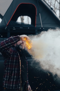 A person wearing a plaid jacket is next to a trail of smoke and sparks, with a rooftop tent mounted on the vehicle visible in the background. The scene is set outdoors, possibly in a camping or survival context.