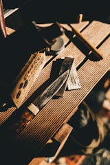 A knife with a polished blade resting on a wooden surface, surrounded by a sharpening stone and a wooden box. Natural light casts shadows, creating a warm and rustic atmosphere.