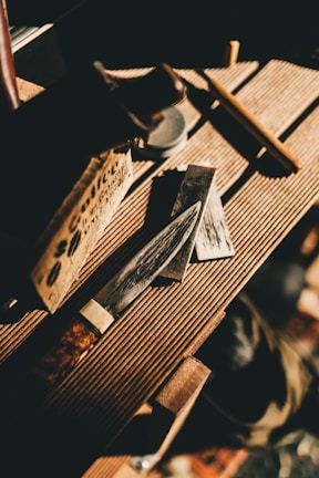A knife with a polished blade resting on a wooden surface, surrounded by a sharpening stone and a wooden box. Natural light casts shadows, creating a warm and rustic atmosphere.