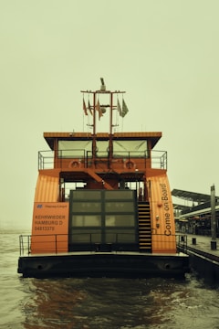 An orange vessel labeled 'Hapag-Lloyd' is docked on a body of water. The vessel features the text 'Come On Board' on its side and has windows, railings, and a staircase leading to an upper deck. Flags are displayed atop the structure. The background portrays an overcast sky and part of a pier.