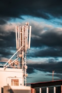 A large telecommunications tower with multiple antennas and equipment is positioned on a rooftop. The sky is filled with dark, dramatic clouds and hints of blue. The sunlight casts warm highlights on the structure, enhancing its metallic components.
