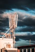 A large telecommunications tower with multiple antennas and equipment is positioned on a rooftop. The sky is filled with dark, dramatic clouds and hints of blue. The sunlight casts warm highlights on the structure, enhancing its metallic components.