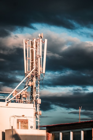 A large telecommunications tower with multiple antennas and equipment is positioned on a rooftop. The sky is filled with dark, dramatic clouds and hints of blue. The sunlight casts warm highlights on the structure, enhancing its metallic components.