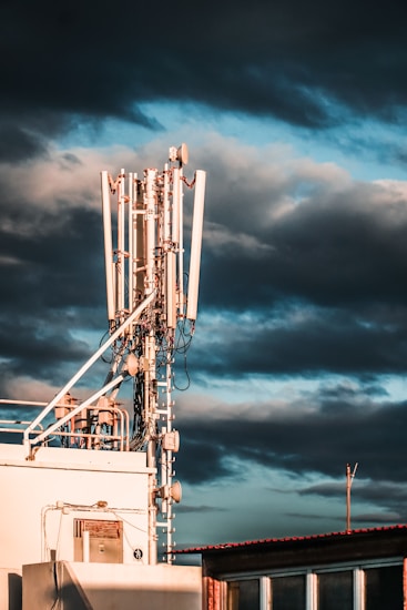 A large telecommunications tower with multiple antennas and equipment is positioned on a rooftop. The sky is filled with dark, dramatic clouds and hints of blue. The sunlight casts warm highlights on the structure, enhancing its metallic components.