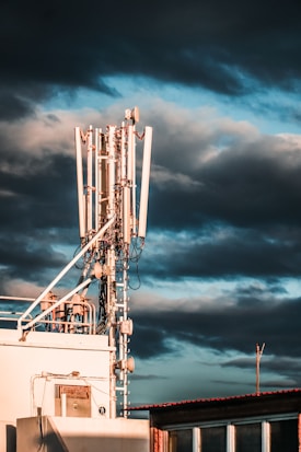 A large telecommunications tower with multiple antennas and equipment is positioned on a rooftop. The sky is filled with dark, dramatic clouds and hints of blue. The sunlight casts warm highlights on the structure, enhancing its metallic components.