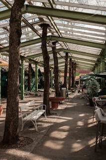 A walkway covered by a green metal and glass pergola with tree trunks integrated into the structure. There are benches, tables, and potted plants along the path, creating a blend of nature and architecture.