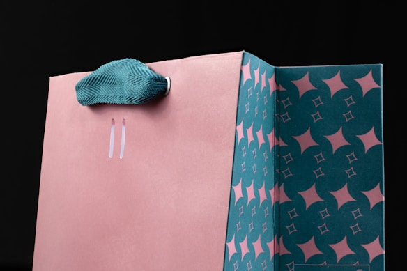 A stylish shopping bag with a salmon pink exterior and a teal handle. The interior features a geometric pattern in teal and pink with star-like shapes. The bag appears to be made of sturdy paper or cardboard.
