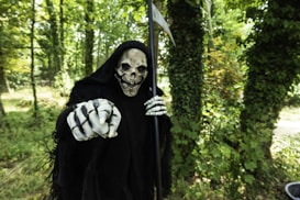 A figure dressed as the Grim Reaper stands in a forest, pointing towards the camera. The figure wears a black cloak and a skull mask, with bony hands prominently visible. Green trees and foliage fill the background, creating a contrast with the dark attire of the figure.
