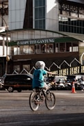 A child wearing a helmet rides a bicycle in an urban setting. The background features a busy street with several cars and motorcycles. In the backdrop, there is a building with a sign reading 'Kopi Titi Gantung', and an orange traffic cone is visible nearby.