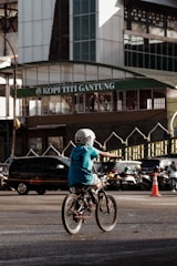 A child wearing a helmet rides a bicycle in an urban setting. The background features a busy street with several cars and motorcycles. In the backdrop, there is a building with a sign reading 'Kopi Titi Gantung', and an orange traffic cone is visible nearby.