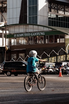A child wearing a helmet rides a bicycle in an urban setting. The background features a busy street with several cars and motorcycles. In the backdrop, there is a building with a sign reading 'Kopi Titi Gantung', and an orange traffic cone is visible nearby.