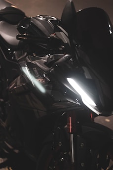 A close-up view of a sleek, modern motorcycle with a glossy black finish. The motorcycle features sharp lines and a prominent headlight, reflecting light in a dimly lit environment. Metallic and black components blend seamlessly, giving it a futuristic and powerful appearance.