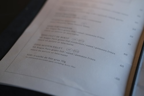 A close-up of a menu page showing various food items with descriptions and prices. The menu lists dishes like SA Angus Pure grass-fed fillet, tenderloin, salad, pommes frites, and foie gras. Some items include abbreviations like GF and DFC, likely indicating dietary information.