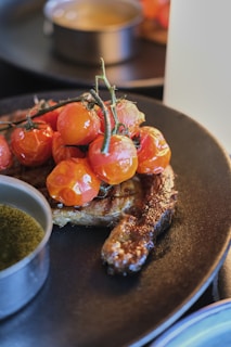 A grilled piece of meat is adorned with a cluster of vine-ripened cherry tomatoes on a black plate. A small dish with a greenish sauce accompanies the dish, and the focus is on the rich texture of the steak and glossy tomatoes. The background is softly blurred, lending a gourmet appeal.