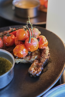 A grilled piece of meat is adorned with a cluster of vine-ripened cherry tomatoes on a black plate. A small dish with a greenish sauce accompanies the dish, and the focus is on the rich texture of the steak and glossy tomatoes. The background is softly blurred, lending a gourmet appeal.