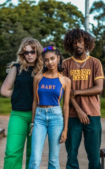 Three young individuals stand together outdoors, with a background of green trees and a clear sky. The person on the left wears dark sunglasses, a black sleeveless top, and bright green pants. The person in the middle has colorful sunglasses on their head and wears a blue sleeveless top with the word 'BABY' in orange, paired with light blue jeans. The person on the right wears a brown T-shirt with 'DADDY COOL' printed in yellow and dark green pants.
