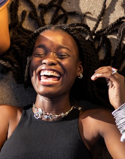 A person is laughing joyfully while lying on the ground. The hair is styled in long braids, and there is a bright, wide smile. The person is wearing a black sleeveless top and layered necklaces. Sunlight creates a warm glow across the face.