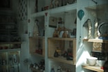 A shop window displaying various artistic and decorative items such as sculptures, pottery, and crafts. The shelves hold abstract sculptures, ceramic figures, a hot air balloon decoration, and a few decorative plates. The overall arrangement is eclectic, with a mixture of both modern and traditional artistic styles.