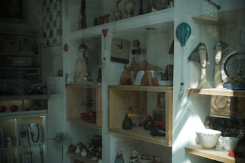 A shop window displaying various artistic and decorative items such as sculptures, pottery, and crafts. The shelves hold abstract sculptures, ceramic figures, a hot air balloon decoration, and a few decorative plates. The overall arrangement is eclectic, with a mixture of both modern and traditional artistic styles.
