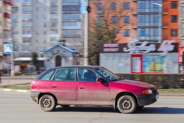 A red compact hatchback car is driving on a city street, with motion blur indicating speed. In the background, there are modern multi-story buildings, a small chapel with a blue roof, and a sign with decorative text.