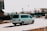 A white van is driving on a city street during daytime. The background includes several parked cars, a billboard, and blurred images of buildings and trees. The van is moving at a noticeable speed as the motion blur suggests.