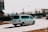 A white van is driving on a city street during daytime. The background includes several parked cars, a billboard, and blurred images of buildings and trees. The van is moving at a noticeable speed as the motion blur suggests.
