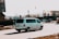 A white van is driving on a city street during daytime. The background includes several parked cars, a billboard, and blurred images of buildings and trees. The van is moving at a noticeable speed as the motion blur suggests.
