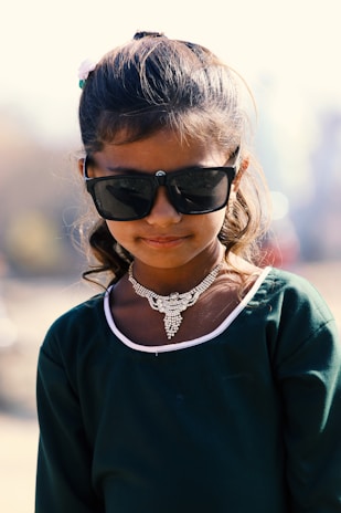 A young girl with dark sunglasses and a decorative silver necklace stands outdoors. She wears a teal dress with white trim and her hair is styled back with a white hair accessory.