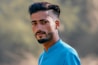 A young man with a neatly groomed beard and hairstyle is wearing a bright blue shirt. He is looking directly at the camera with a neutral expression. The background is blurred, highlighting the subject.