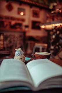 A cozy indoor setting features a cup of coffee beside an open book on a wooden table. In the background, there are blurred images of wall decor and a softly lit interior, suggesting a cafe environment. A small dessert sits on a plate next to the coffee cup.
