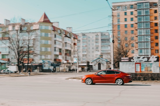 A red sedan is driving on an urban street with several mid-rise apartment buildings in the background. The buildings feature a mix of glass, concrete, and brick materials. The sky is clear, indicating a sunny day. Power lines and a few bare trees are visible.