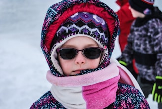 A child is dressed warmly in a patterned winter coat, a knitted hat with geometric designs, and a scarf wrapped around the face. The child is wearing sunglasses, suggesting bright outdoor conditions. In the background, another person is partially visible, wearing a red outer layer.