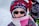 A child is dressed warmly in a patterned winter coat, a knitted hat with geometric designs, and a scarf wrapped around the face. The child is wearing sunglasses, suggesting bright outdoor conditions. In the background, another person is partially visible, wearing a red outer layer.