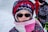 A child is dressed warmly in a patterned winter coat, a knitted hat with geometric designs, and a scarf wrapped around the face. The child is wearing sunglasses, suggesting bright outdoor conditions. In the background, another person is partially visible, wearing a red outer layer.