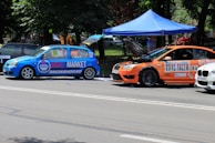 Two modified cars are parked on the side of a road under a blue canopy. The blue car features prominent branding with 'Grill Market' on the side, while the orange car has its hood open, indicating it might be undergoing maintenance or inspection. Both vehicles display various sponsor logos and decals and have racing numbers. The background shows a park-like setting with trees and a few people.